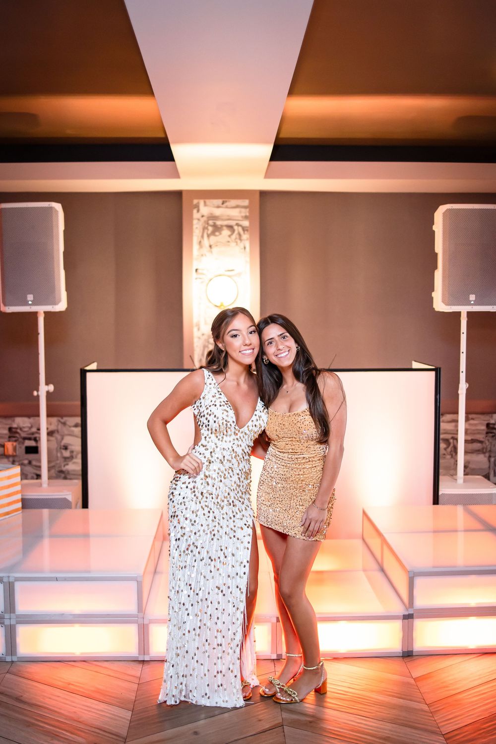 Two women pose together at an event venue wearing sparkly formal dresses with speakers and lighting visible in background.