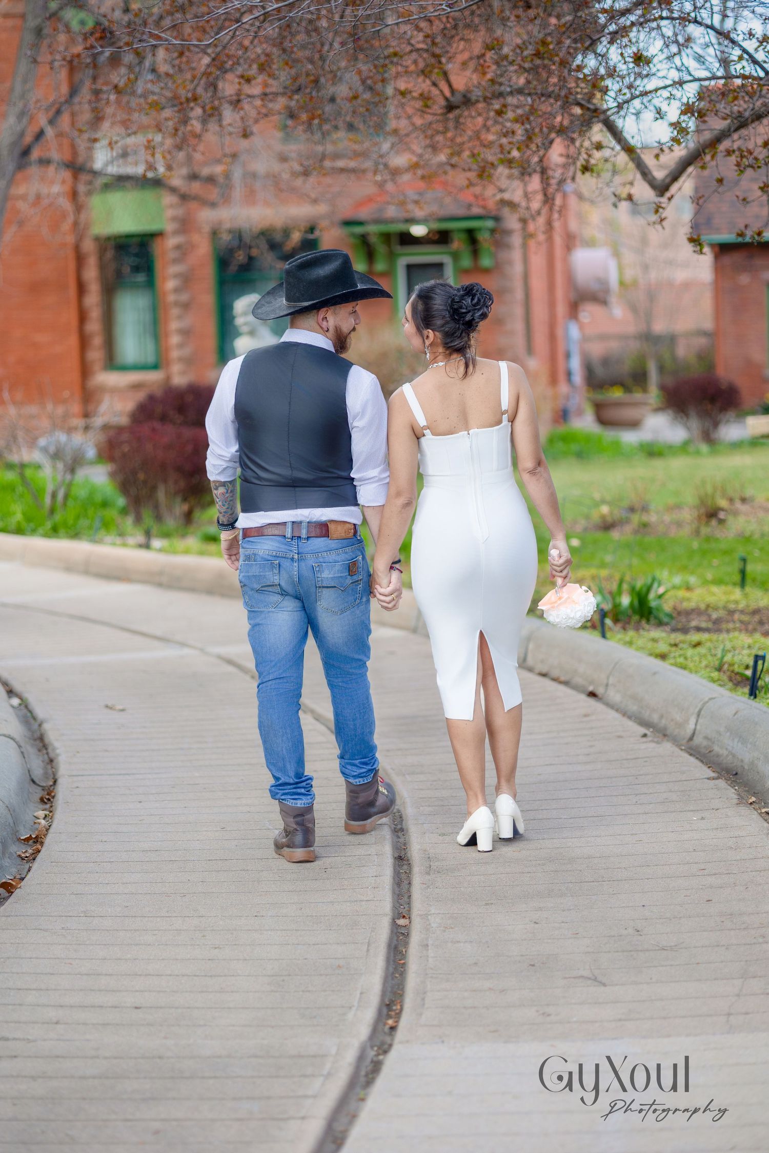 Couple walking together on a sidewalk, with the person in white wearing a dress and the other in denim and vest.
