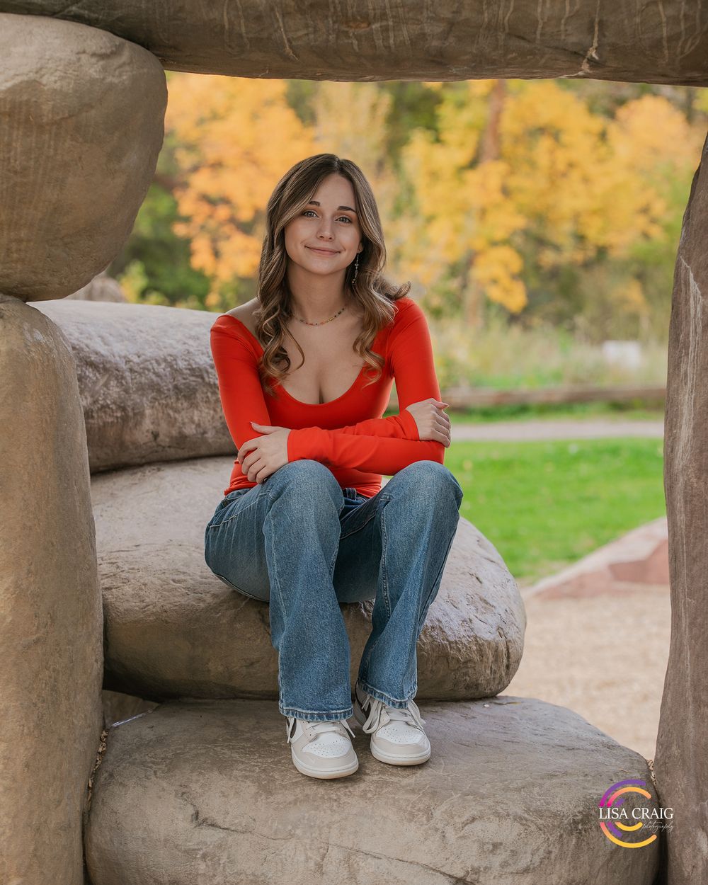Casual outdoor portrait sitting on stone structure with autumn colors in background.