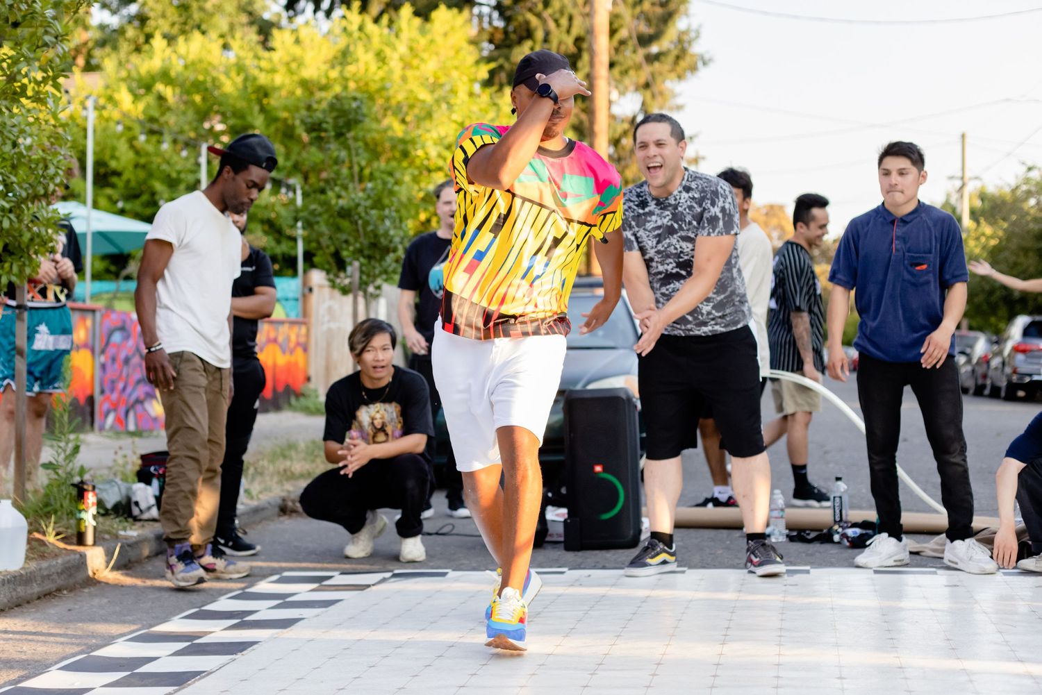 Skateboarder in a yellow shirt performs a trick on a checkered floor at an outdoor skate park while others watch.
