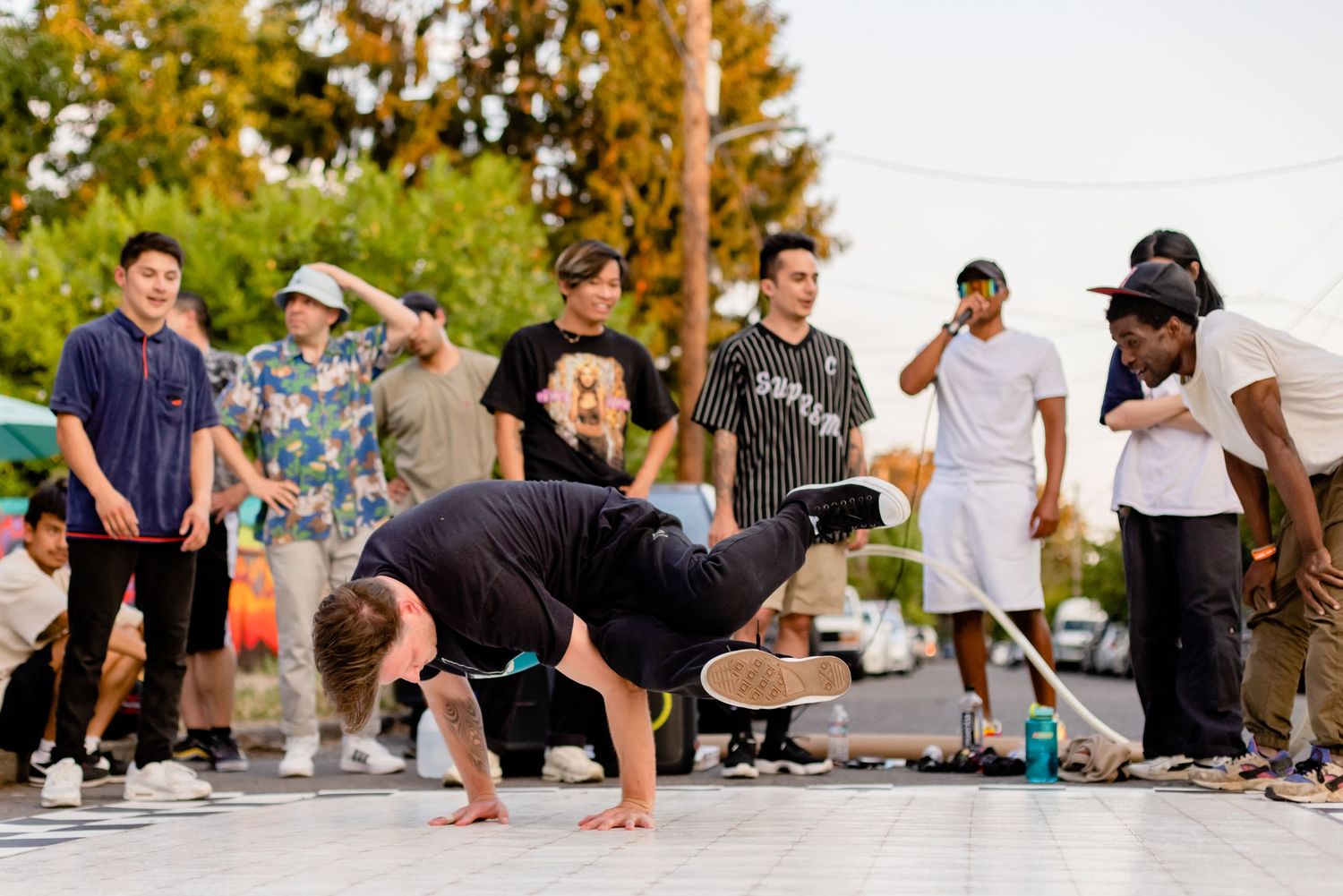 Street dancers perform breakdancing moves on concrete while others watch and cheer in an outdoor setting.