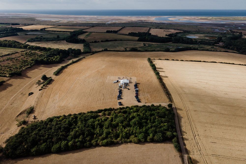 Aerial view of rural farmland with golden fields surrounding a small farm complex, with the sea visible on the horizon.
