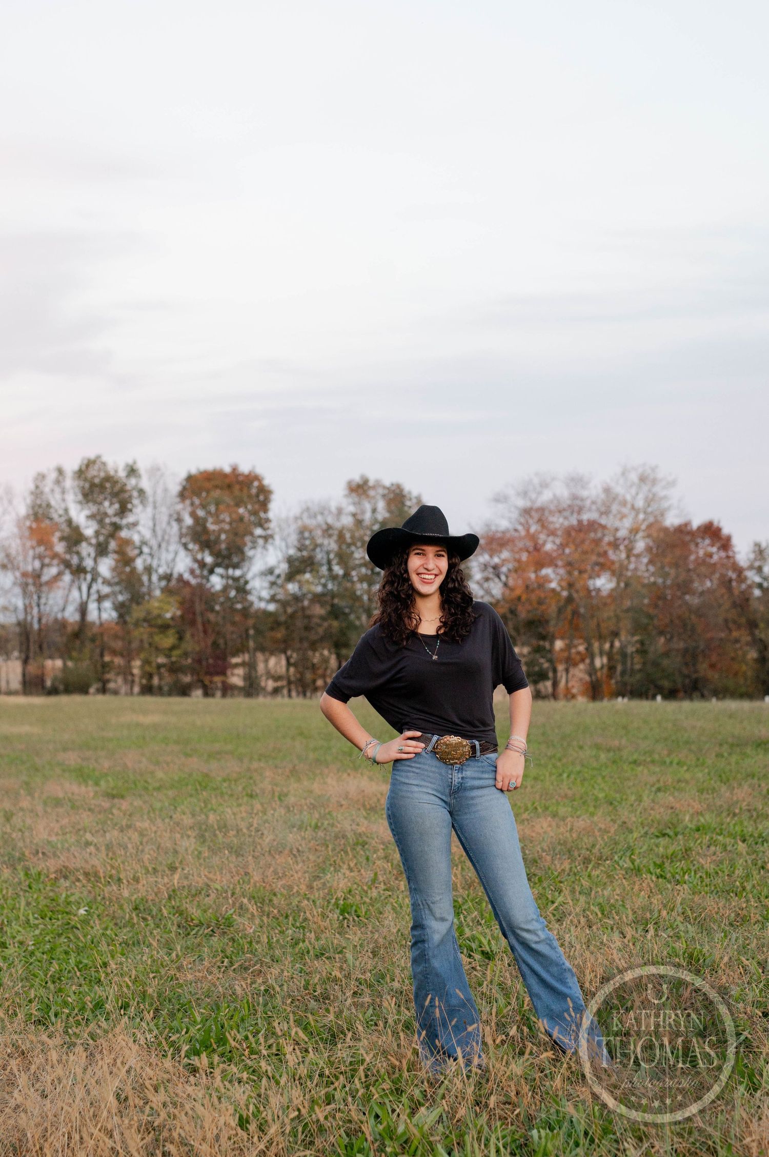 Figure in black top and jeans strikes confident pose in autumn field wearing cowboy hat.