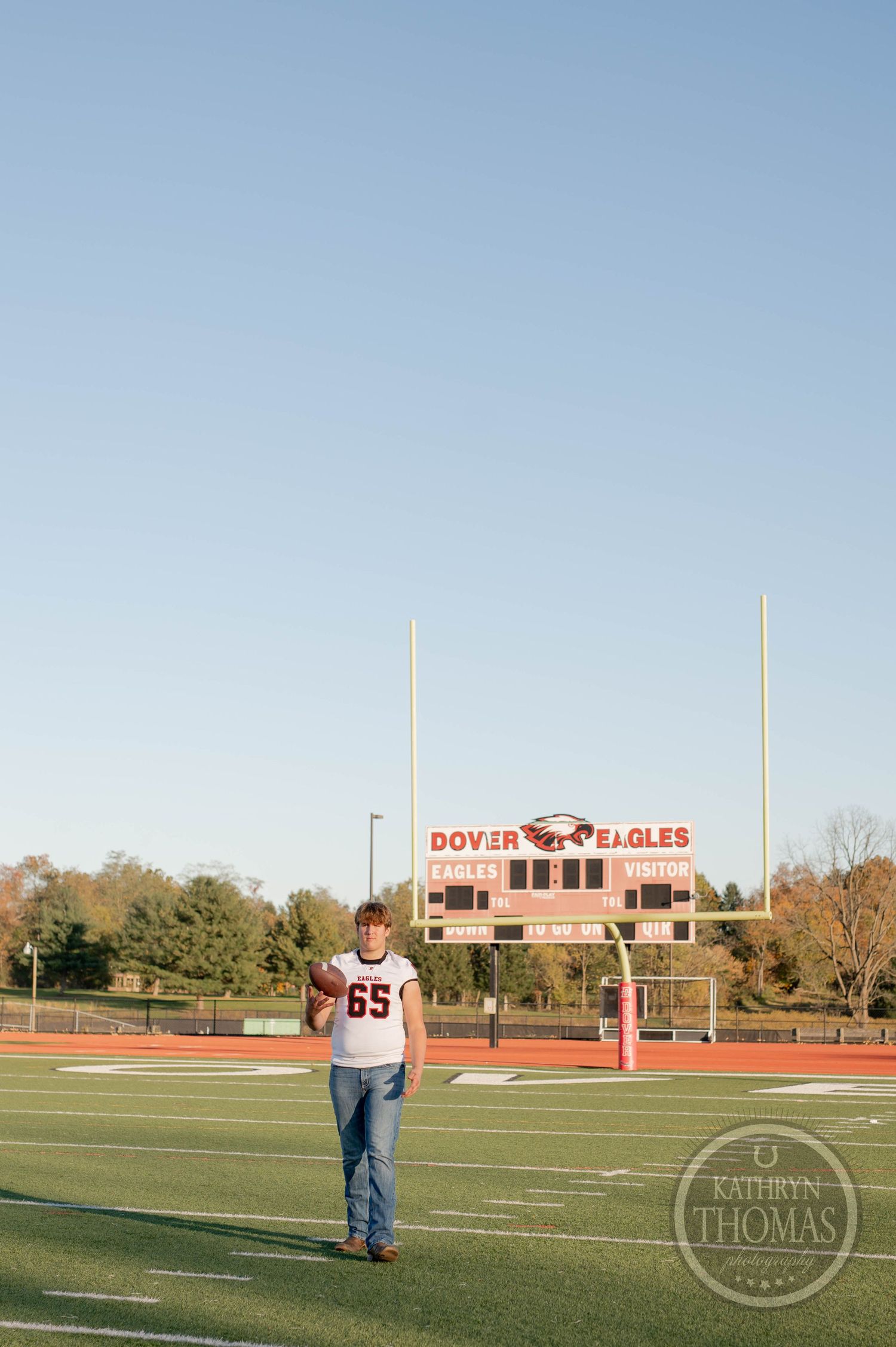 Player in jersey 65 throws football on field with Dover Eagles scoreboard visible in background.