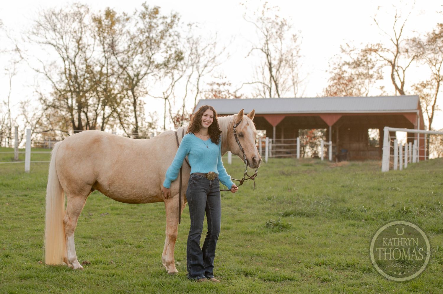 Person in turquoise sweater stands with palomino horse on grassy hill near barn.