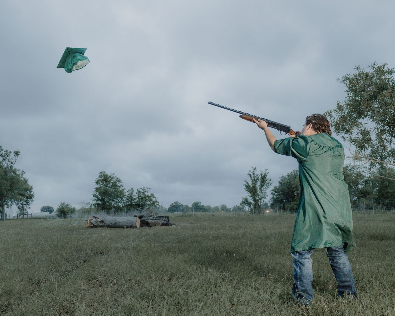 Clay pigeon shooter in green jacket aims shotgun at flying target under cloudy sky in open field.