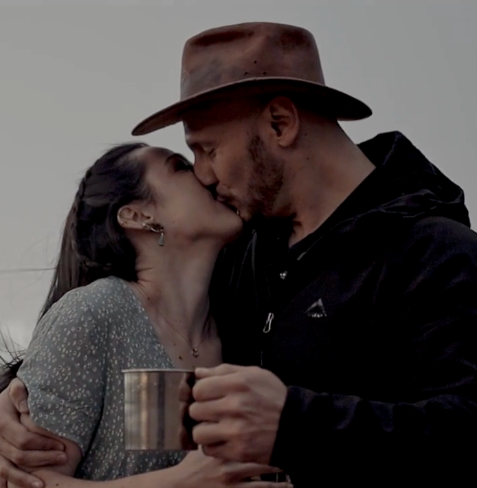 Couple sharing a romantic kiss while holding a coffee cup, with person wearing a brown fedora hat.