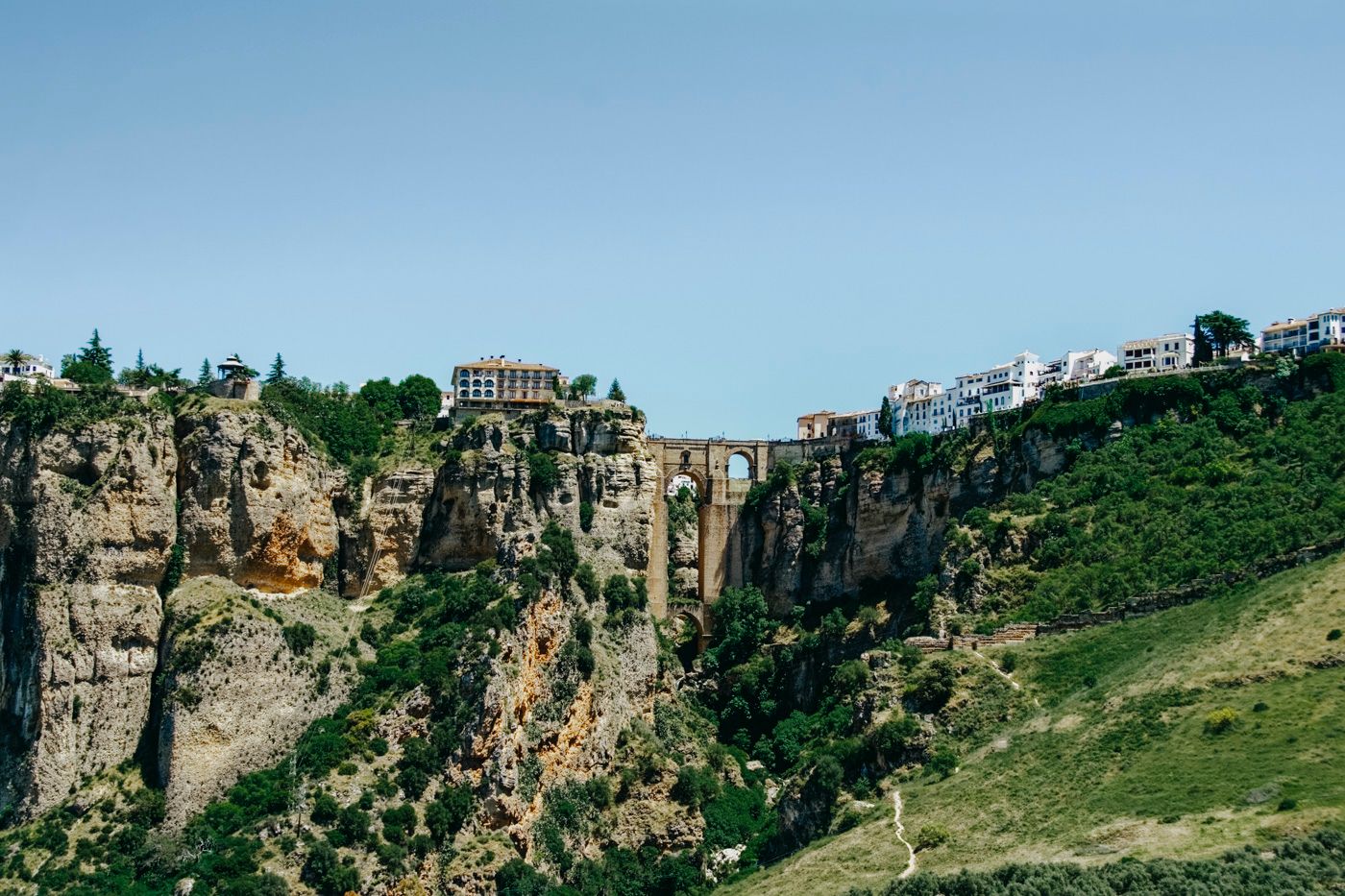 Dramatic cliffside buildings overlook a deep gorge in Ronda, Spain, with blue sky and green hills in view.