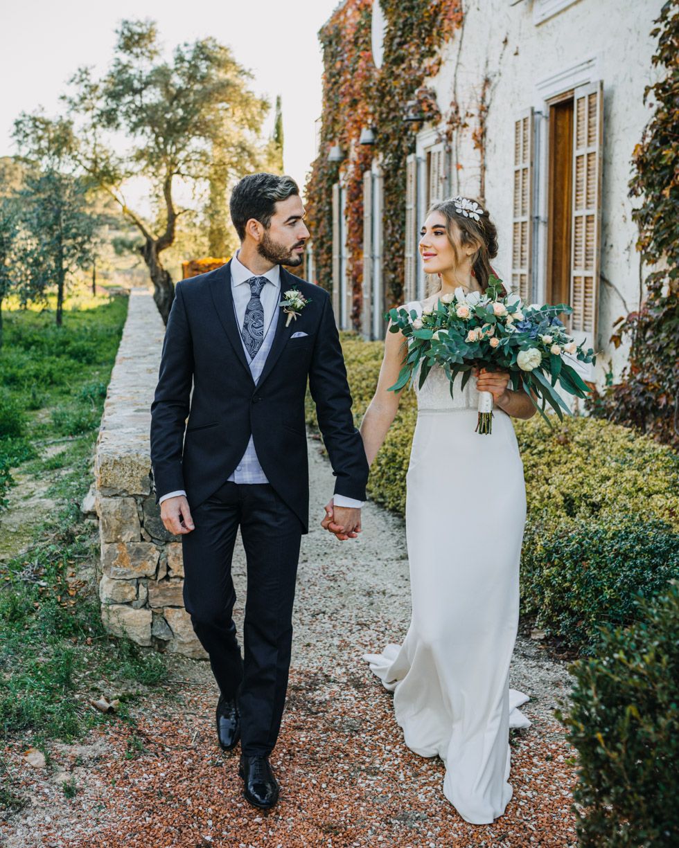 A newlywed couple walks hand in hand along a stone path beside an ivy-covered building on their wedding day.