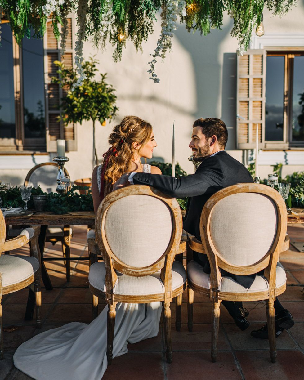 A couple in wedding attire share an intimate moment while seated at elegant upholstered chairs in an outdoor garden setting.