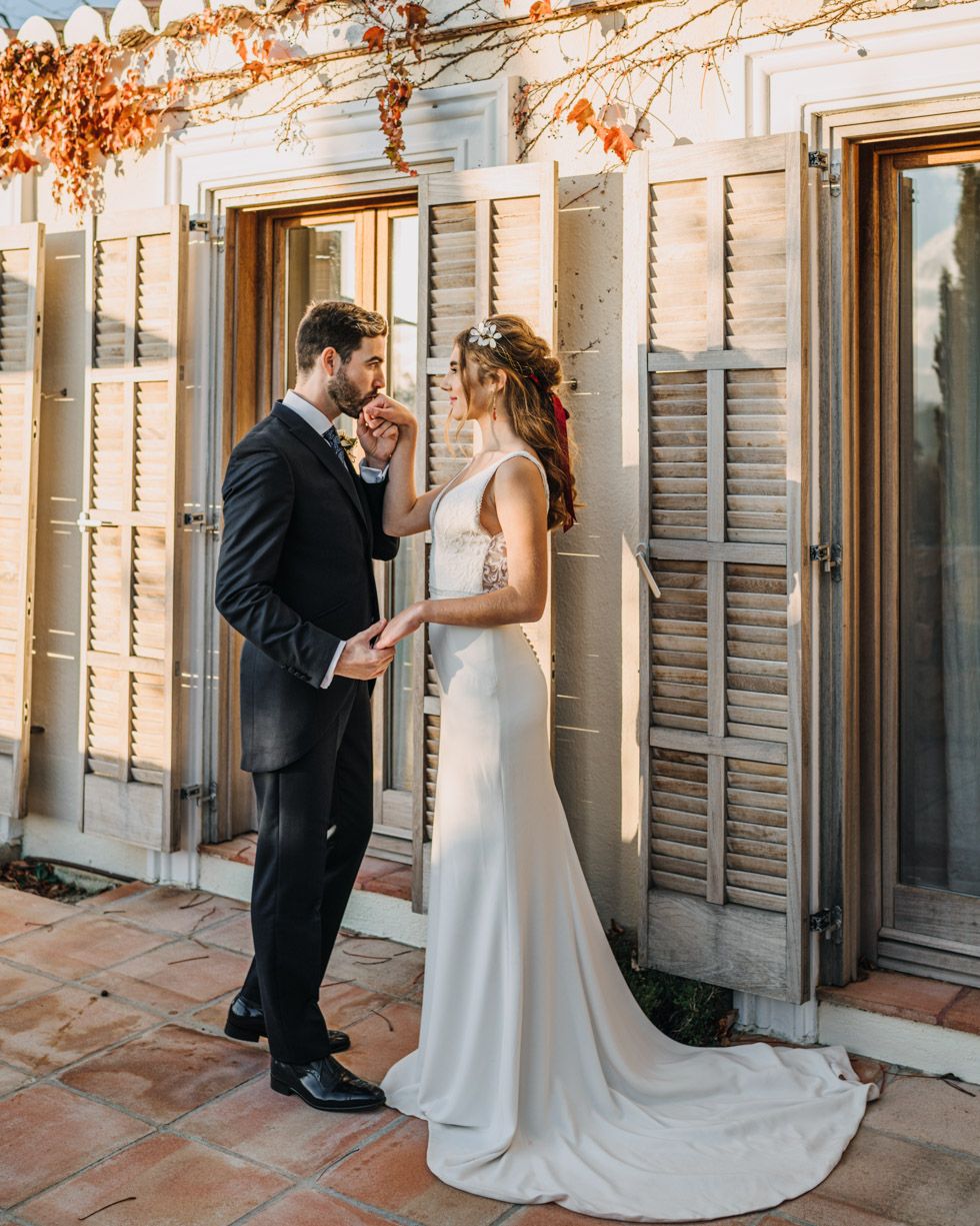 Romantic golden hour wedding portrait of couple in formal attire standing against rustic shuttered building.