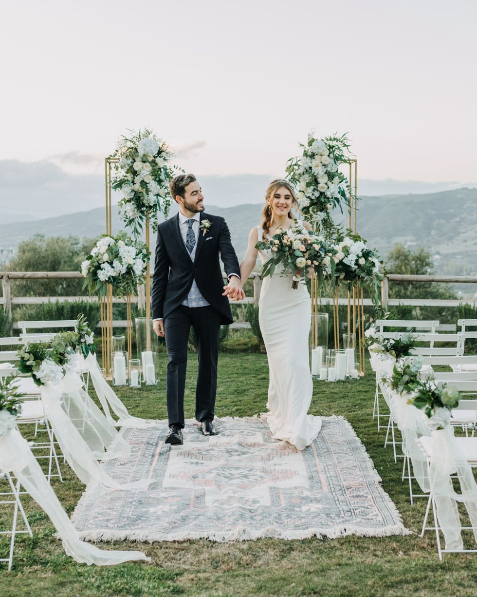 A couple stand together at their outdoor mountain wedding ceremony site with white floral arches and decorative details.