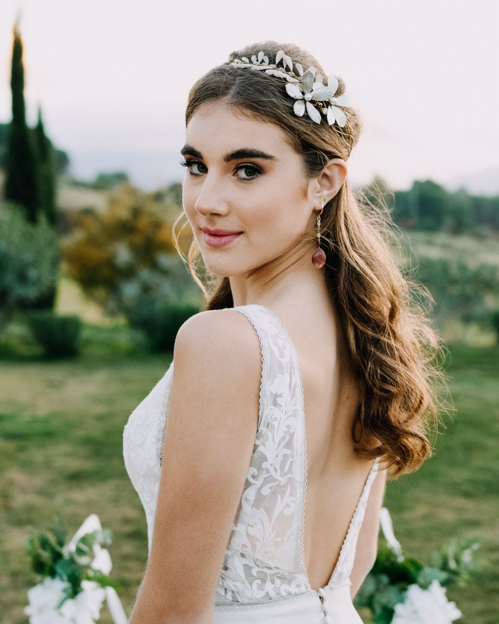 A bride in a white lace wedding dress smiling outdoors with a delicate floral headpiece and flowing curled hair.