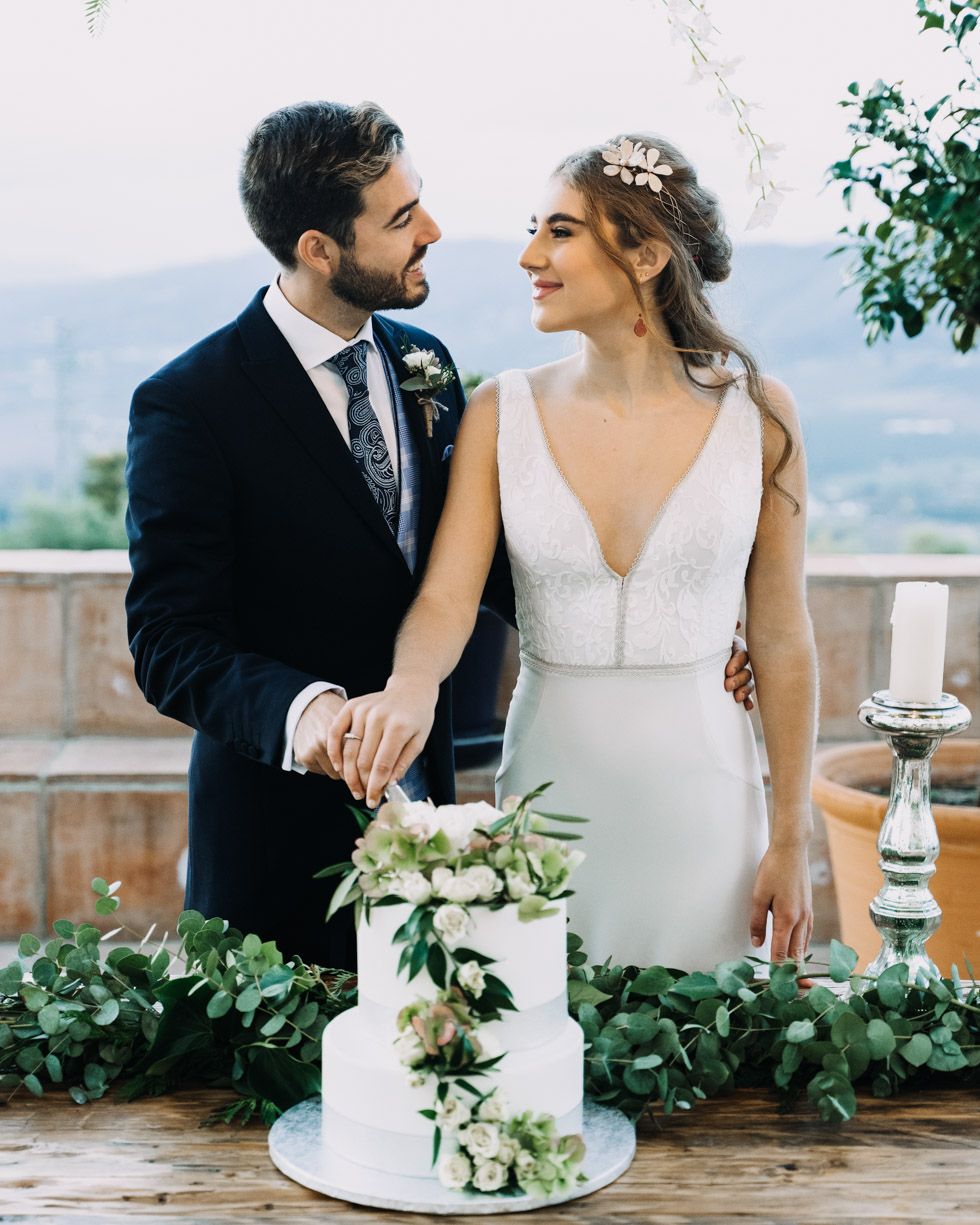 A wedding couple stands next to their elegant white cake adorned with greenery on an outdoor terrace with scenic views.