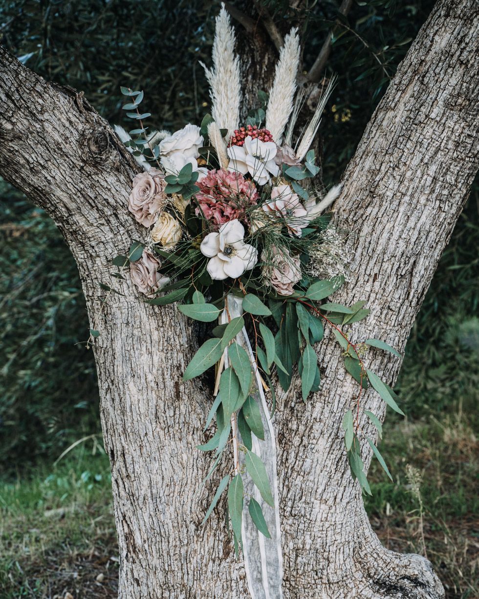 Boho-style floral arrangement with pampas grass and pink roses displayed on a weathered tree trunk in a forest setting.