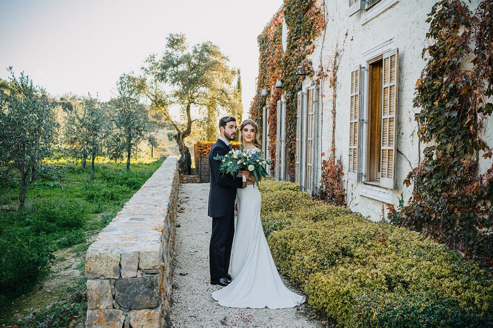 A couple embraces on a stone pathway next to a rustic ivy-covered building during a sunset wedding ceremony.