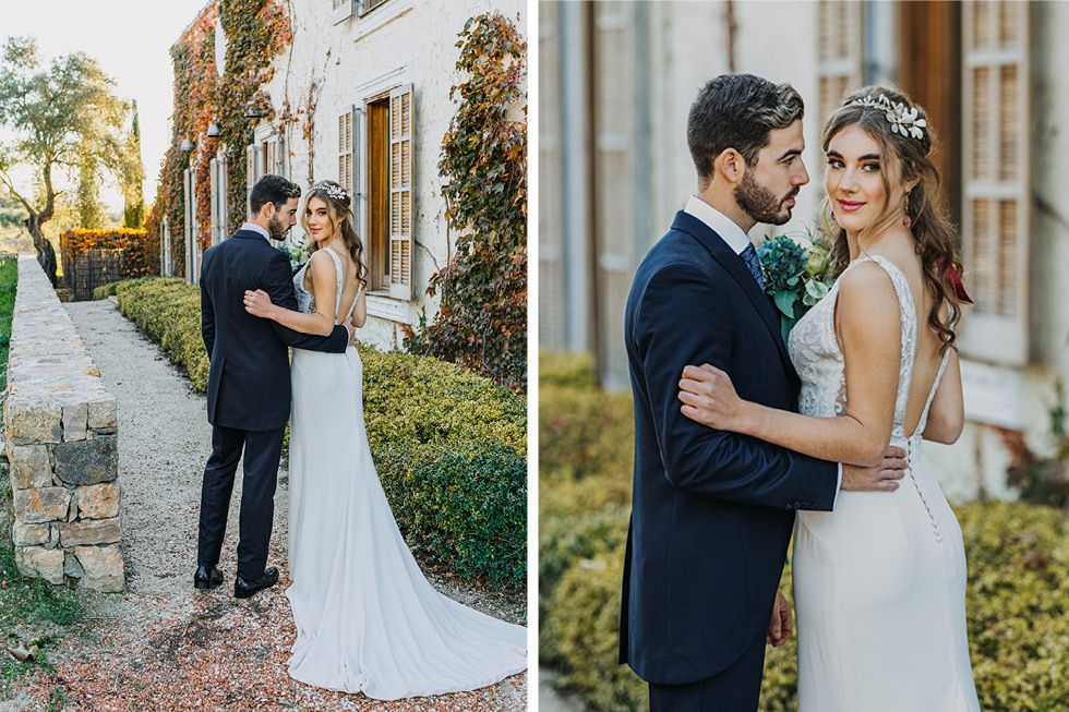 A romantic couple in wedding attire embraces in front of a Mediterranean-style building with lush greenery.