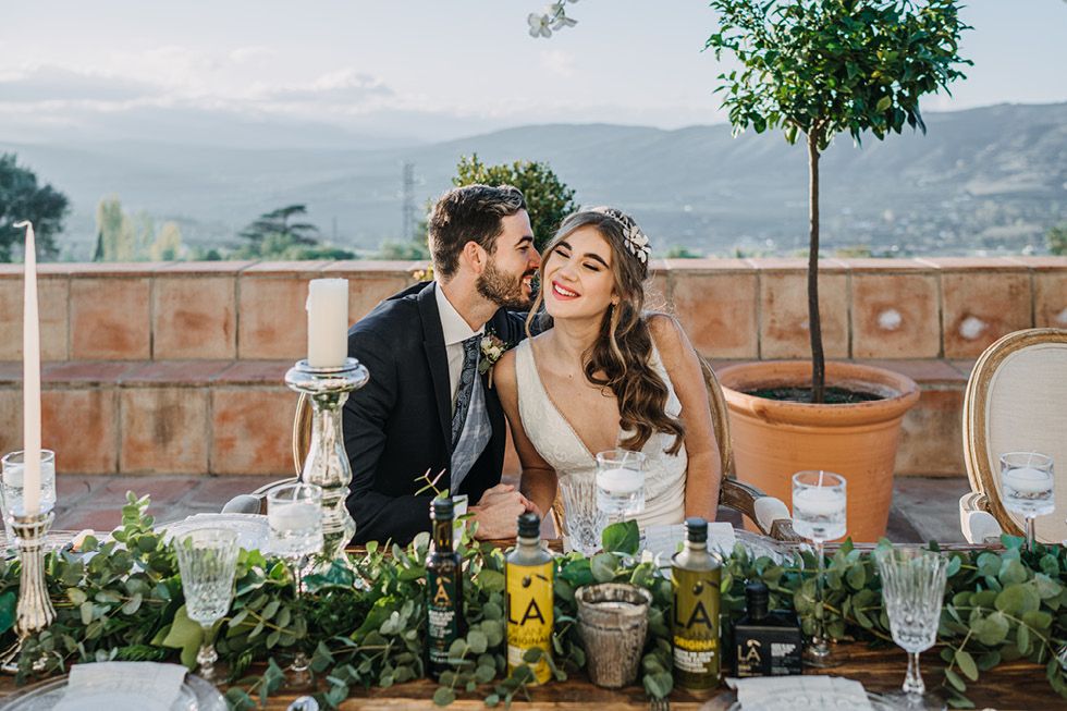 Newlyweds share an intimate moment at an outdoor reception table decorated with floral arrangements and glassware.