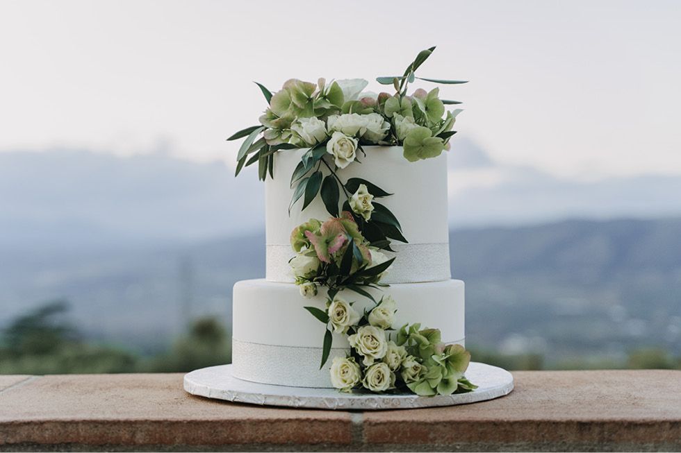 A white two-tier wedding cake decorated with cascading green and white flowers sits outdoors against a mountain backdrop.