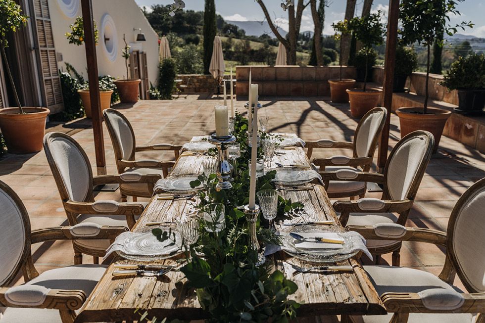 Elegant outdoor dining table set with greenery centerpiece and wooden chairs on sunny Mediterranean terrace.