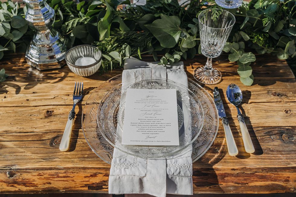 Elegant table setting with clear glass charger plates, menu cards and eucalyptus garland on rustic wooden table.