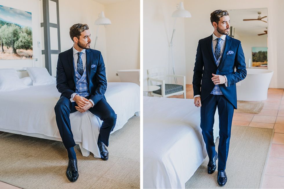 A groom in a navy blue suit with light blue tie sits and stands in a brightly lit white hotel room.