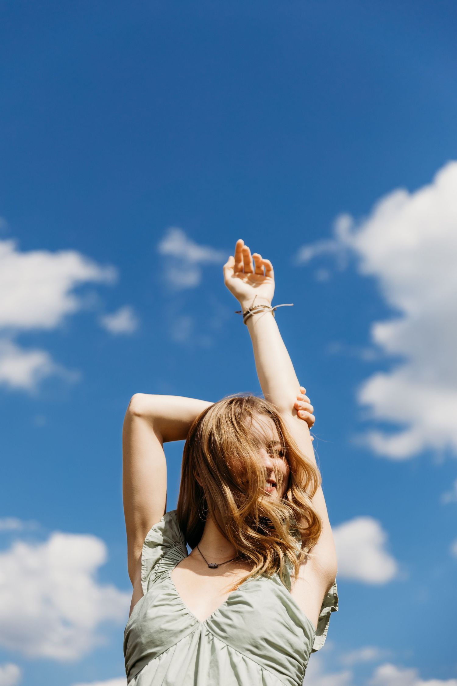 Person in light summer dress stretches joyfully against blue sky with white fluffy clouds on sunny day.