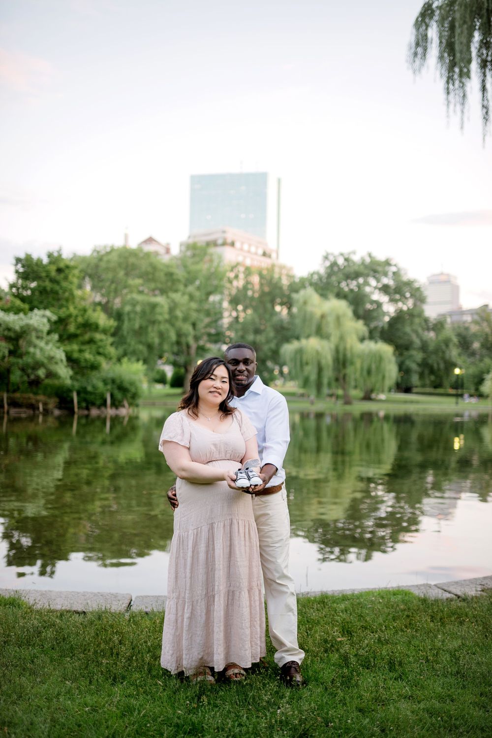 Couple poses by a pond in Boston Public Garden, woman in pink dress holding baby shoes at golden hour.