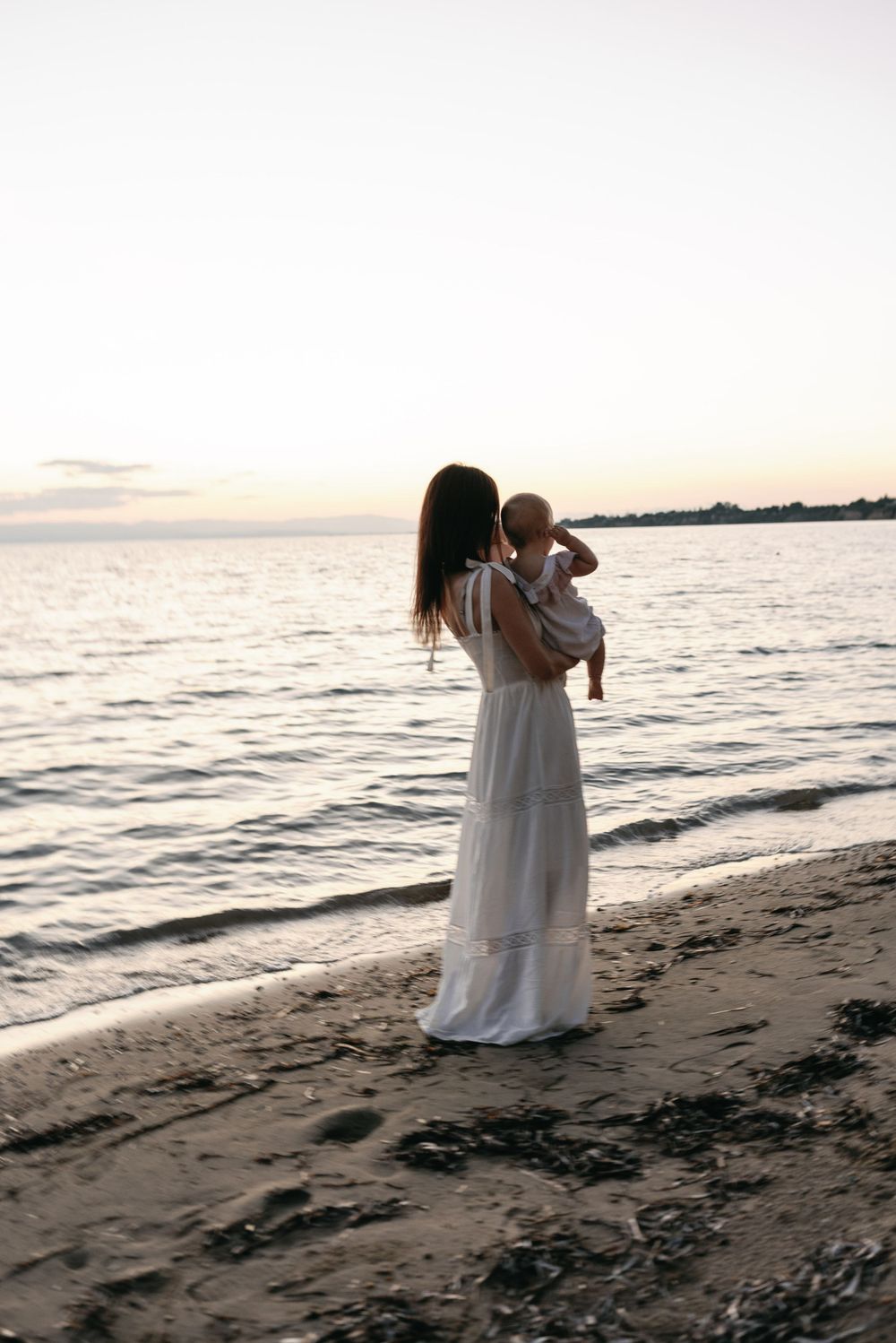 Woman in white dress holds baby while standing on beach at sunset by the ocean's edge.