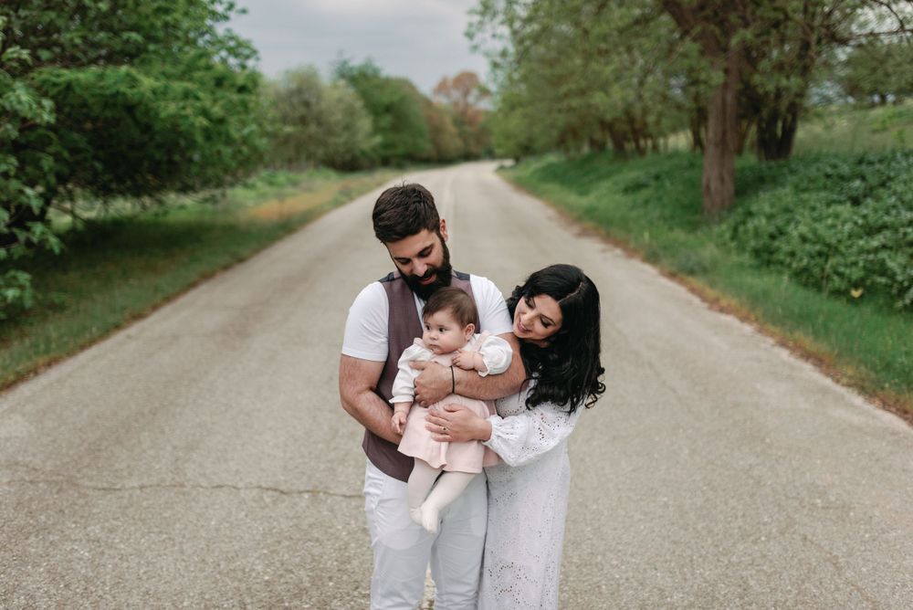 A couple wearing white outfits holds their baby while standing on a peaceful tree-lined gravel path.