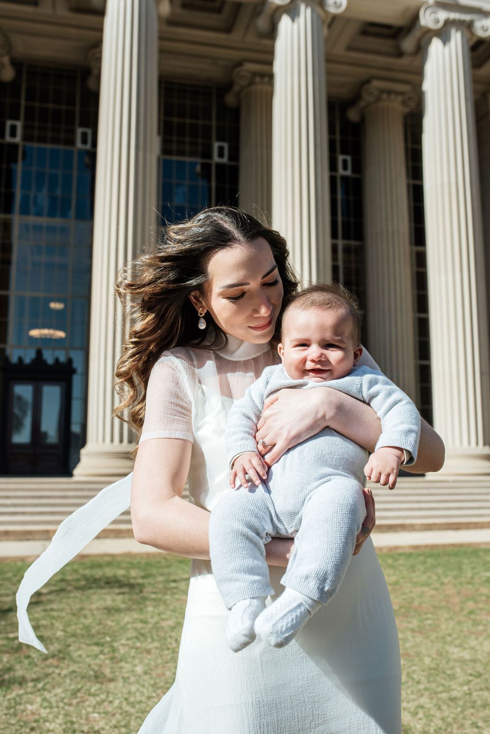 A smiling baby in light blue clothing is held outside a neoclassical building with white columns on a sunny day.
