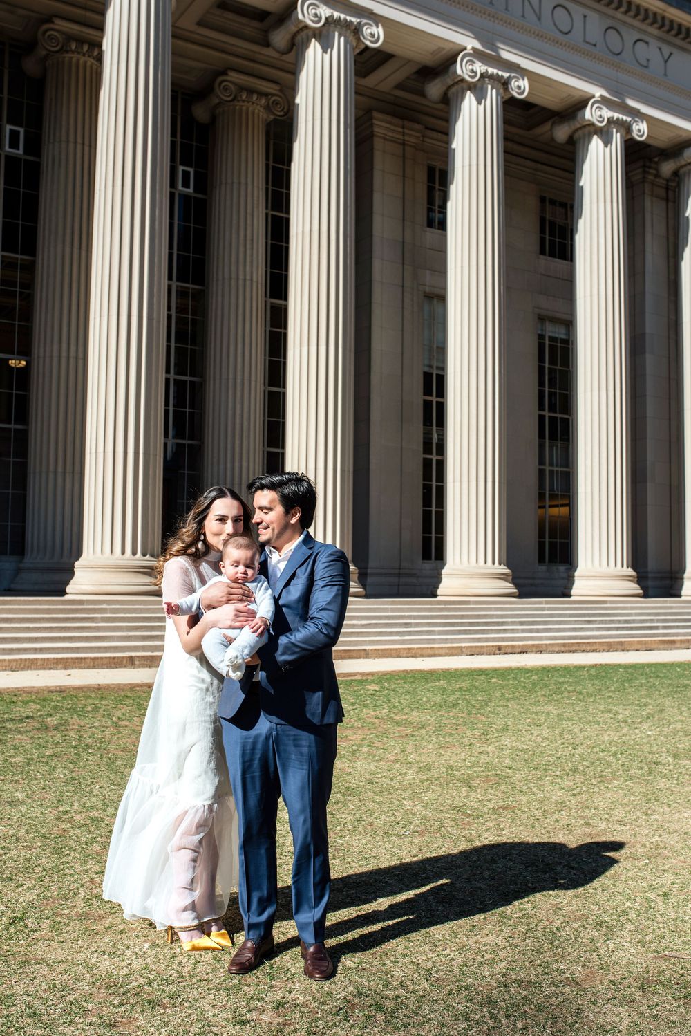 A couple poses with their baby in front of grand classical columns on a grassy lawn at a historic building.