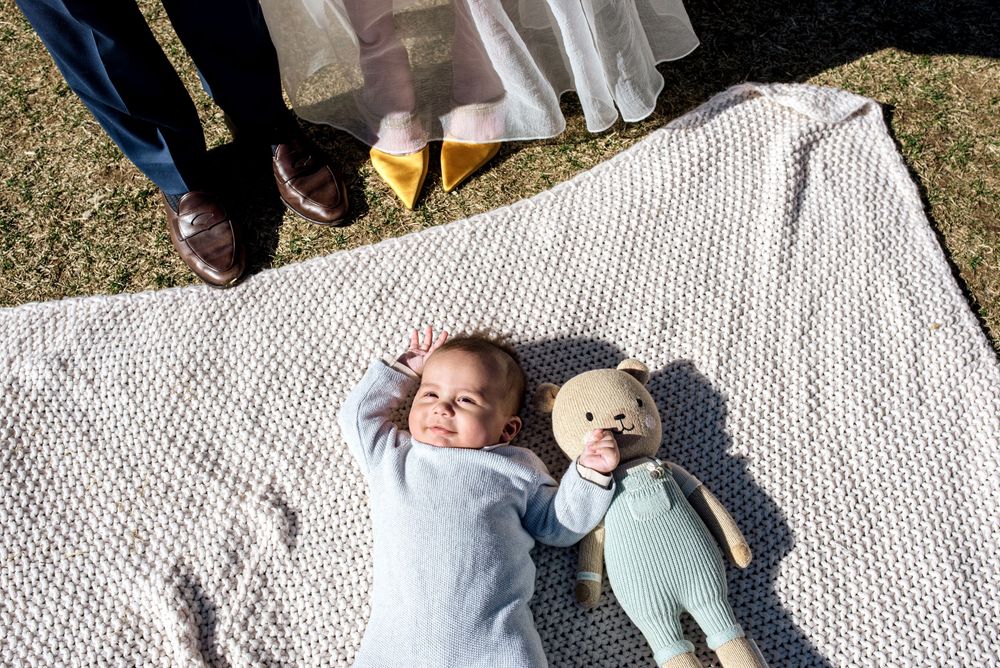 A baby lying on a white blanket outdoors with a teddy bear companion on a sunny day.