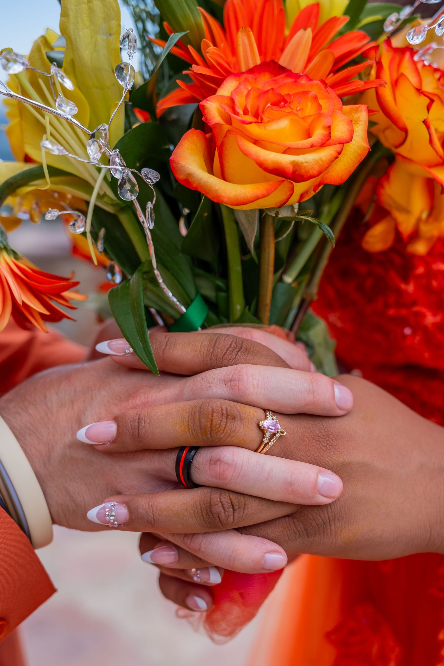 Hands wearing engagement ring and nail art hold vibrant orange roses and flowers against red background.