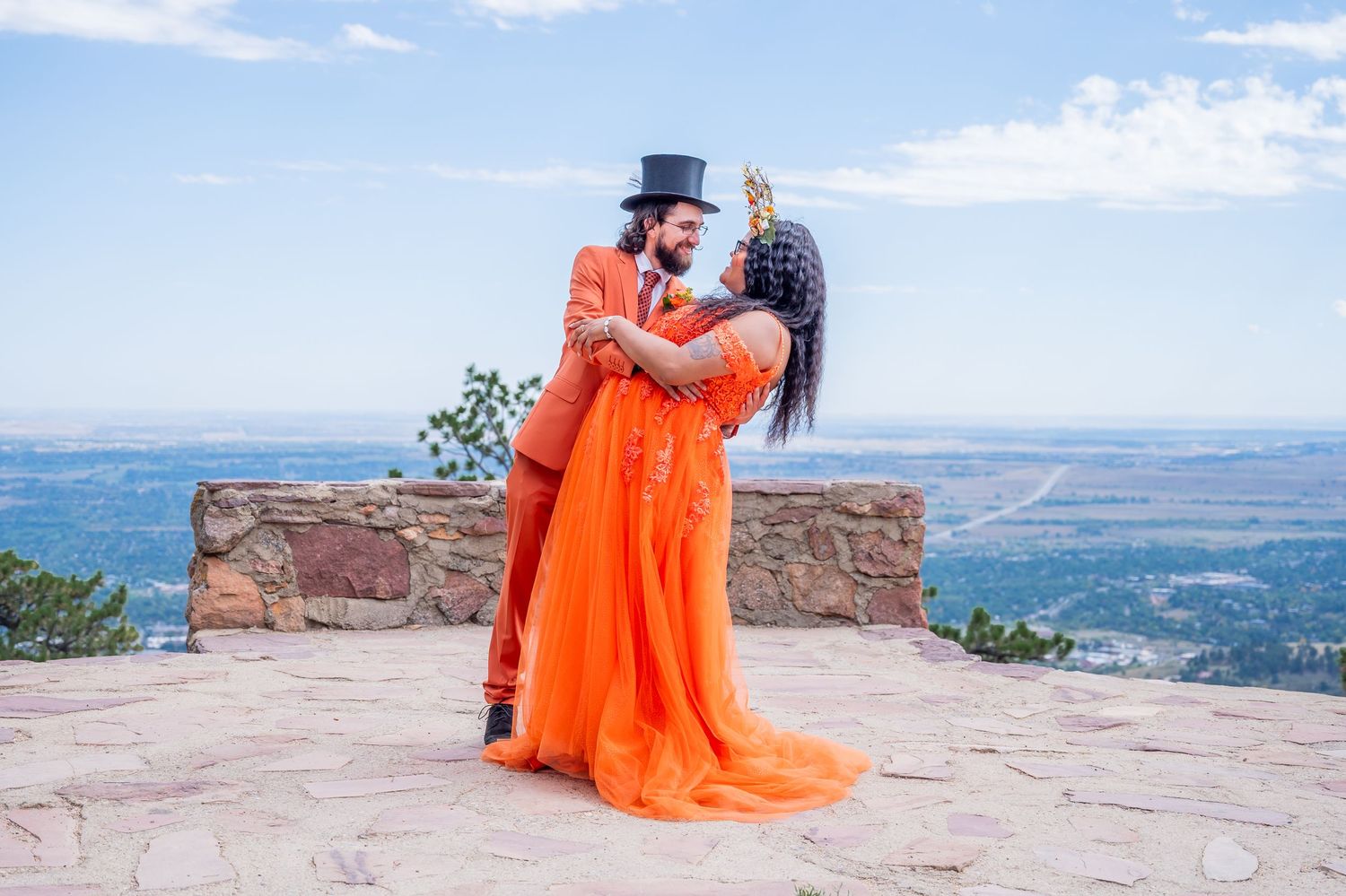 A couple in a formal orange dress and top hat embrace on a stone overlook with mountain views in the background.