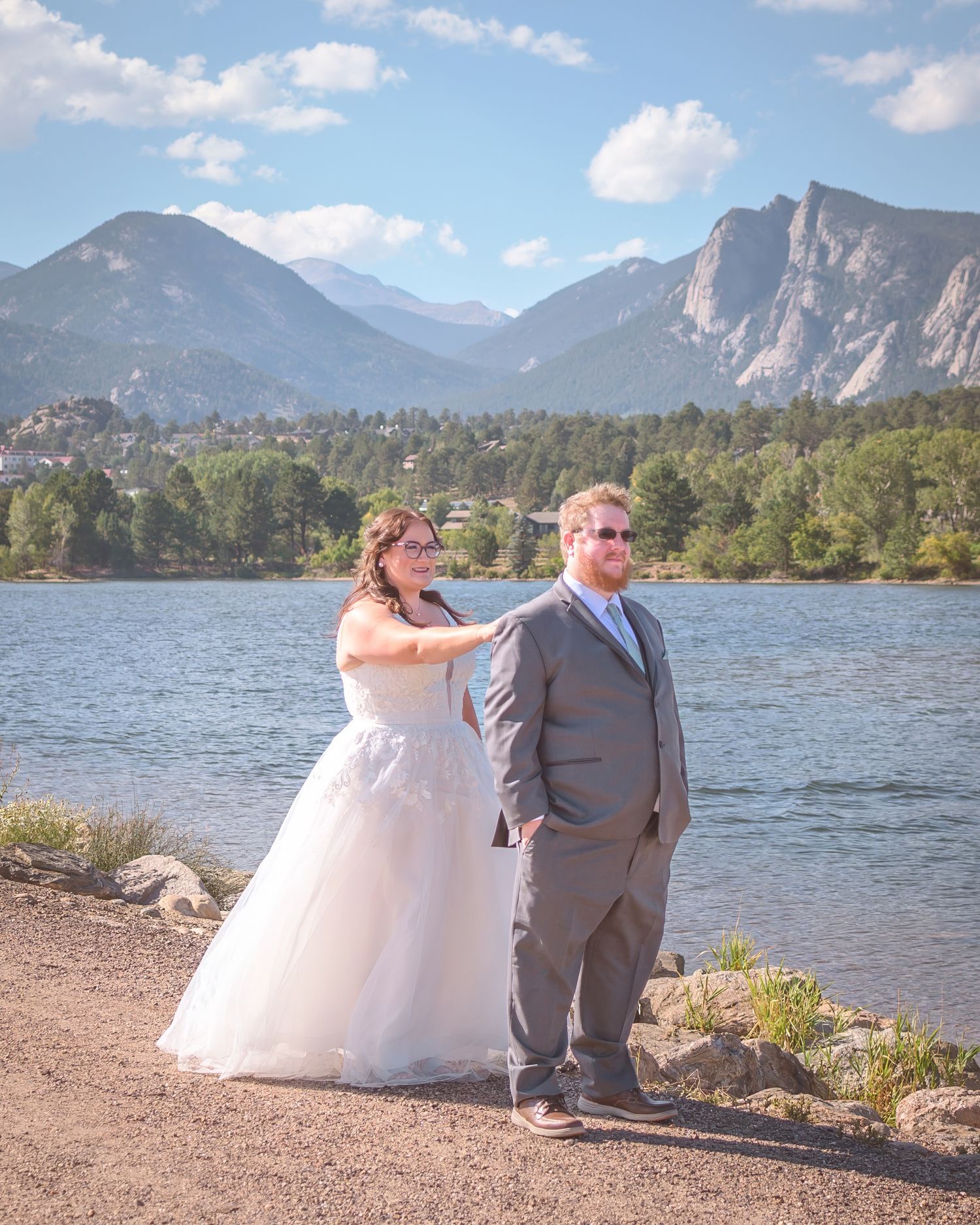 A couple in wedding attire stands by a lake with scenic Rocky Mountain peaks in the background at sunset.