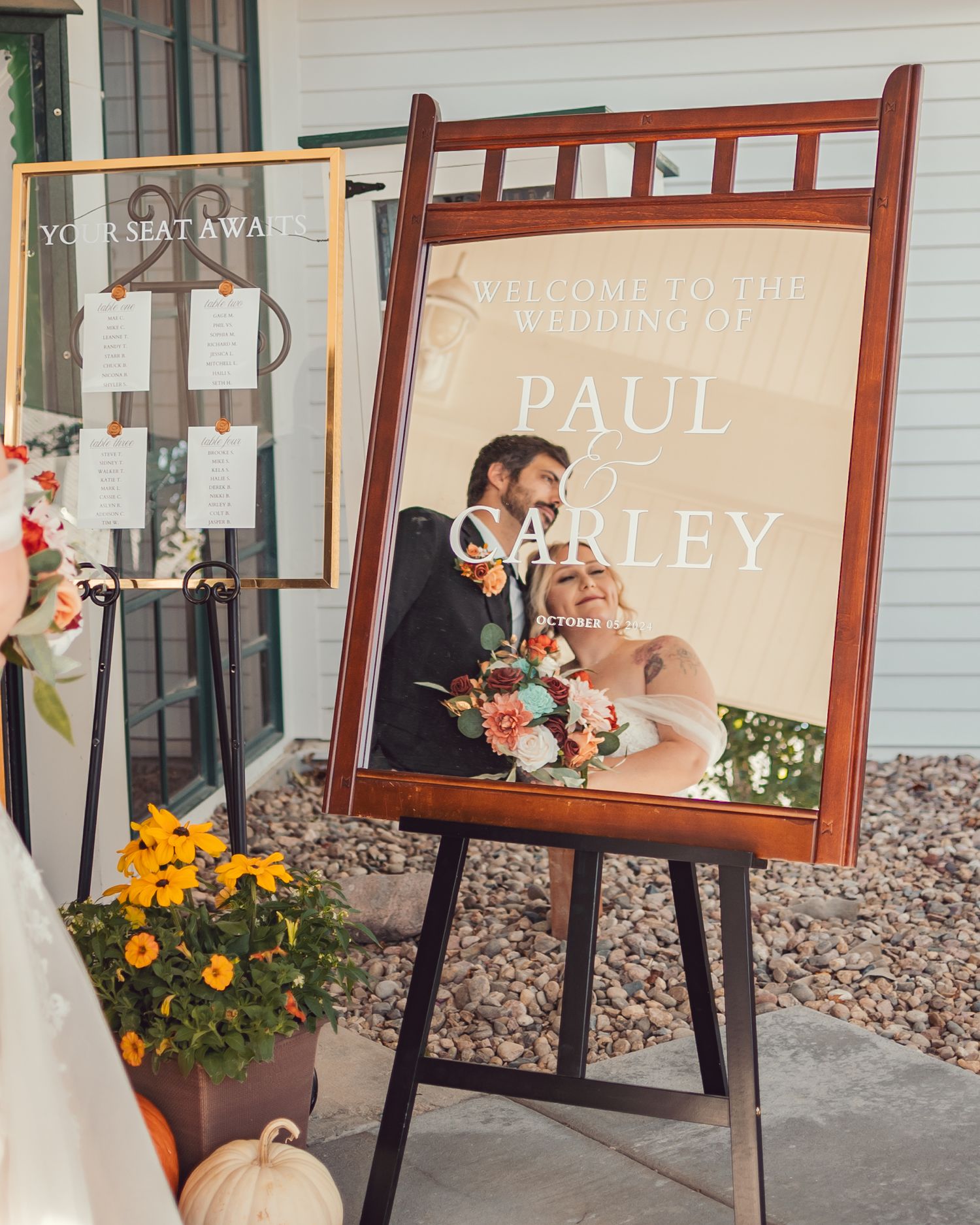 A wooden framed wedding welcome sign on an easel with orange flowers blooming in a planter beside it.