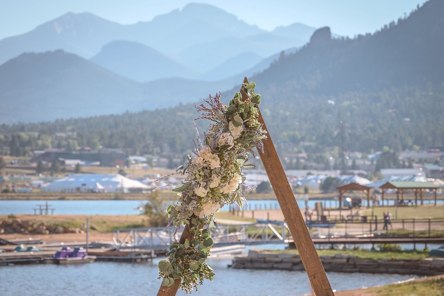 A wooden wedding arch decorated with white flowers overlooks a scenic mountain lake and harbor view.