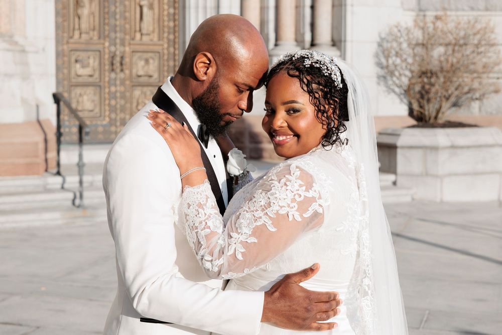 Groom tenderly embracing bride in lace long-sleeve wedding gown with veil outside stone cathedral.