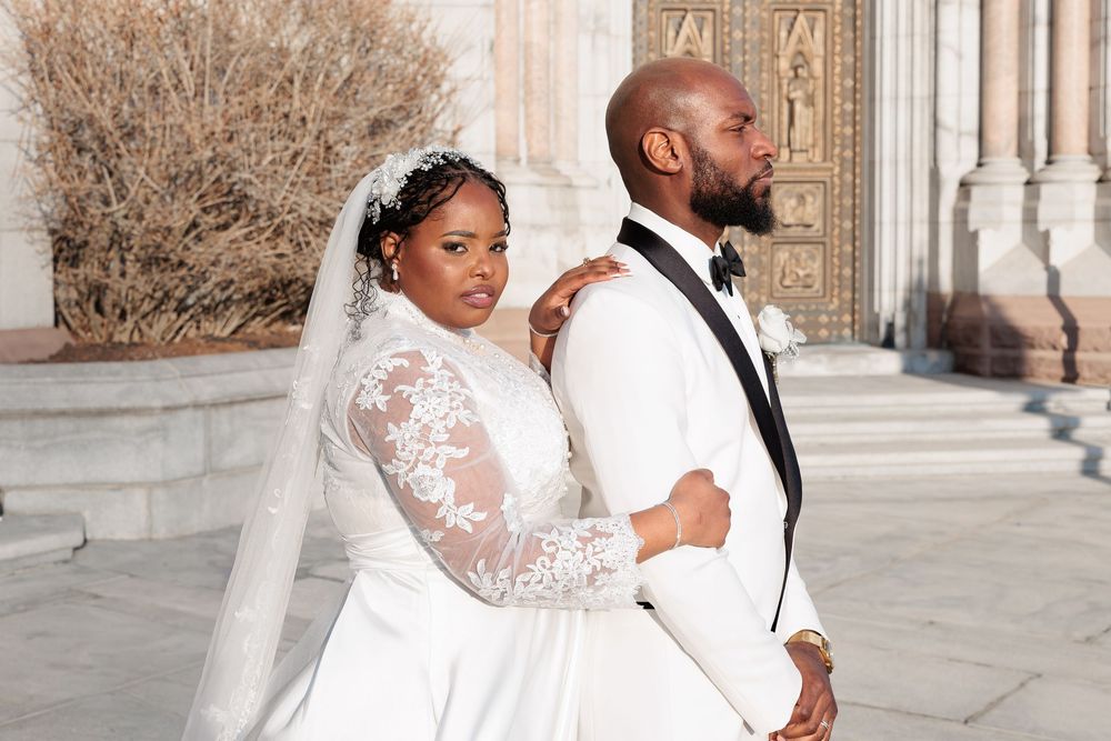 Wedding couple posing back-to-back outside a stone church, bride in lace gown with cathedral veil.
