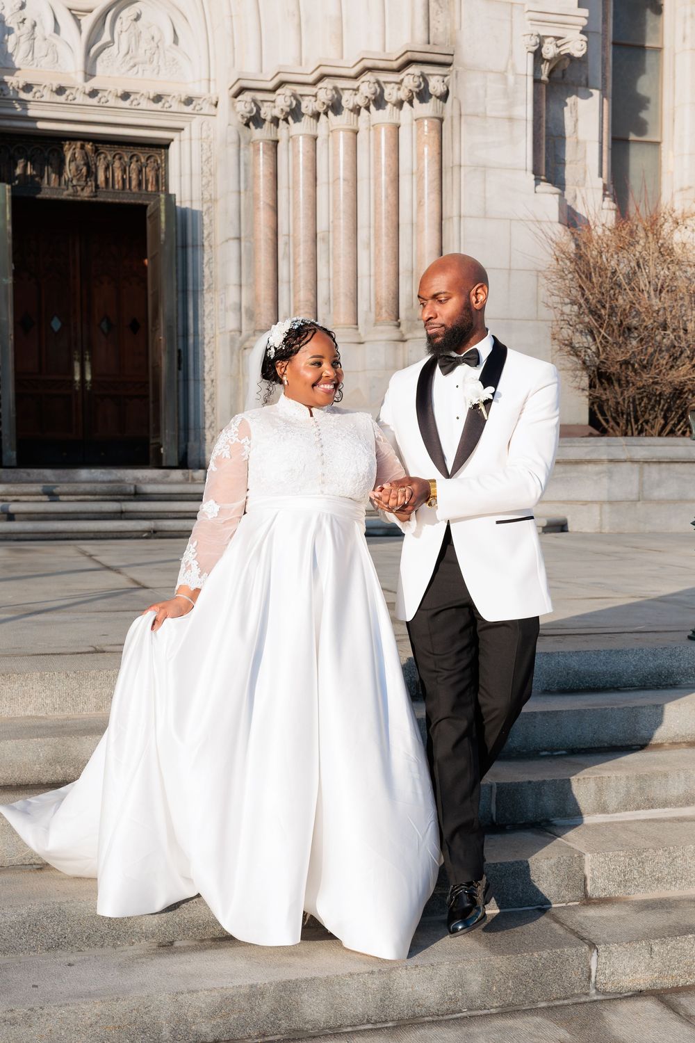 A Black bride and groom hold hands on church steps, she in a white gown, he in a white tuxedo jacket.