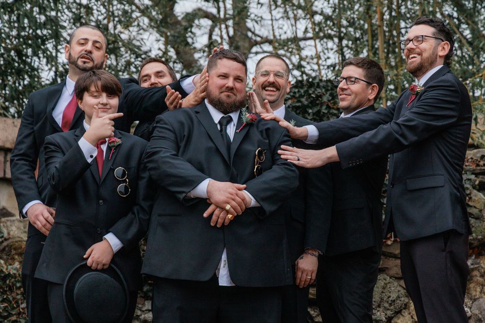 A groom stands proudly with his groomsmen in dark suits, sharing a fun outdoor group moment.