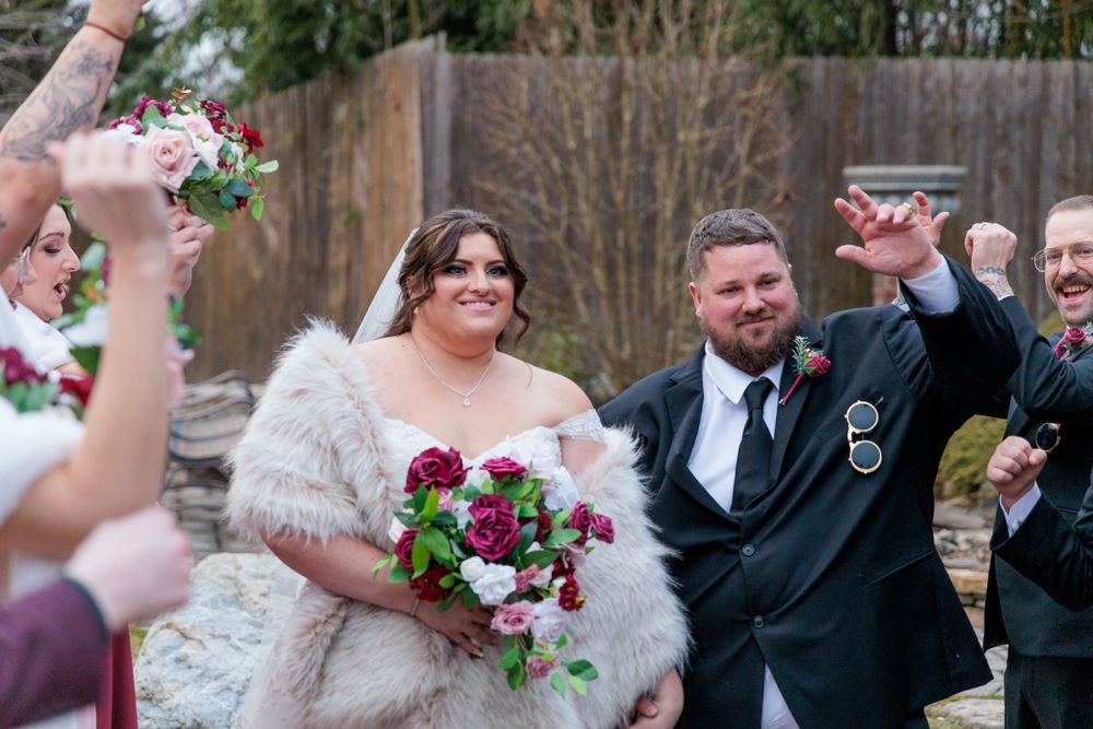 Happy bride and groom exit ceremony as guests cheer, bride carries red and pink rose bouquet.