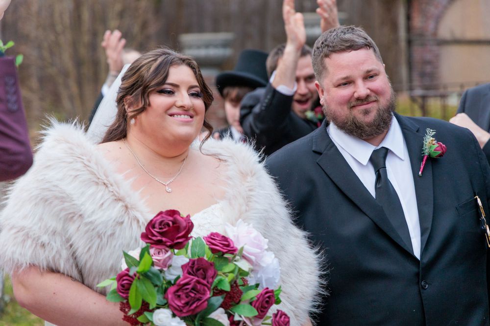 Joyful newlyweds walk down the aisle, bride holding burgundy bouquet in fur stole, guests celebrating behind them.