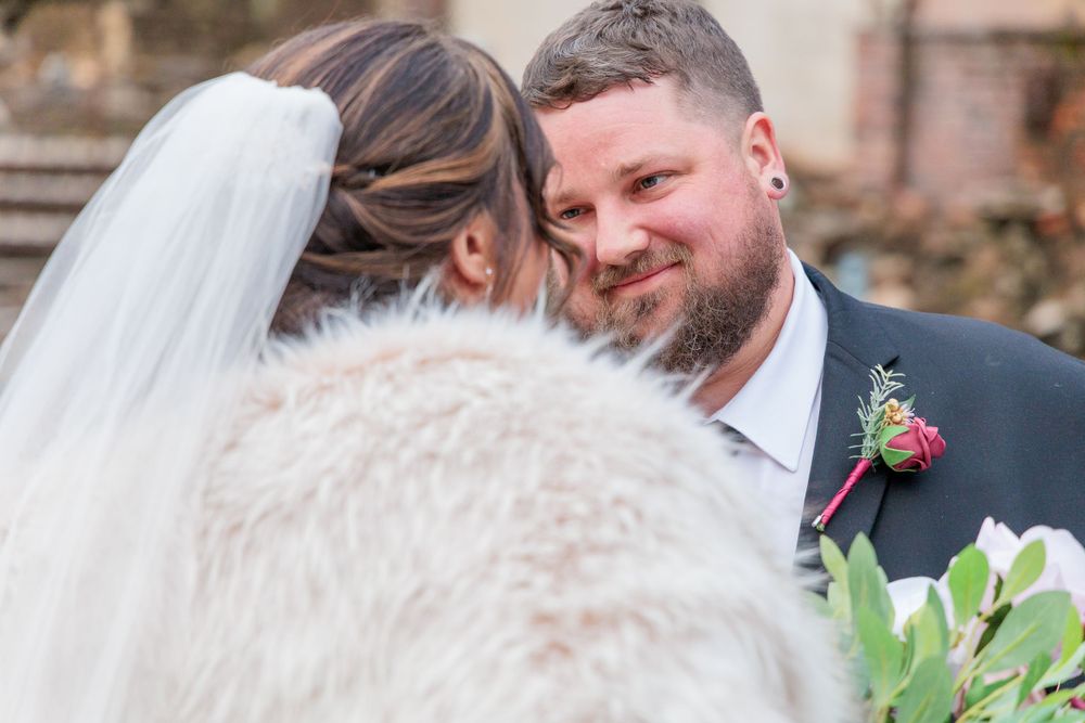 Groom smiles tenderly at bride wearing fur wrap during intimate wedding ceremony moment outdoors.