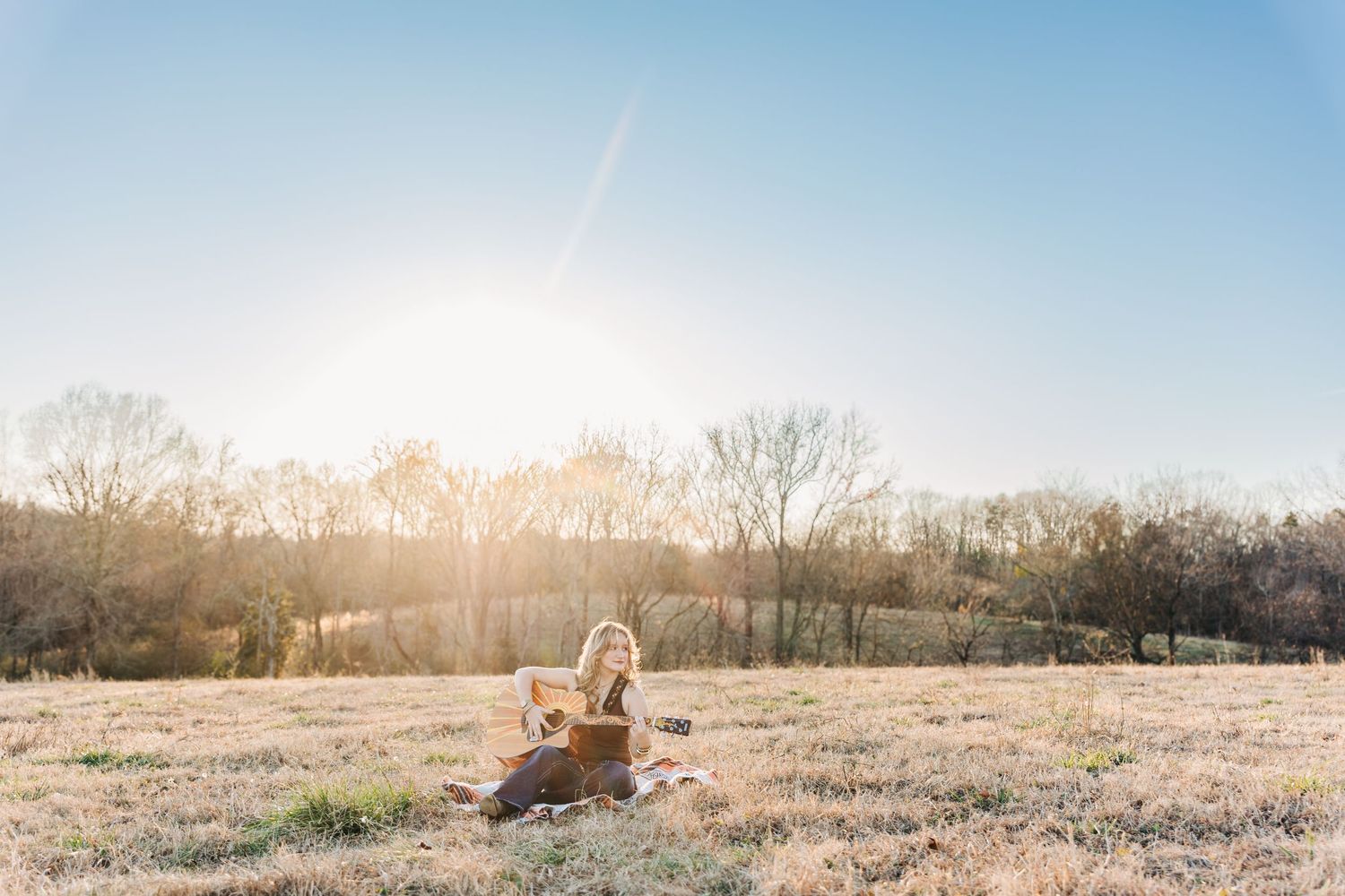 A panoramic sequence of golden sunset light illuminating a grassy field with trees along the horizon with a young woman playing guitar.