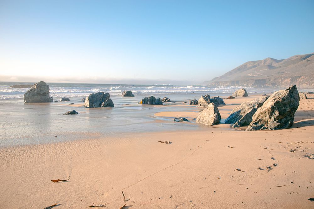 A panoramic view of a sandy beach with rock formations and mountains in the distance under a blue sky.