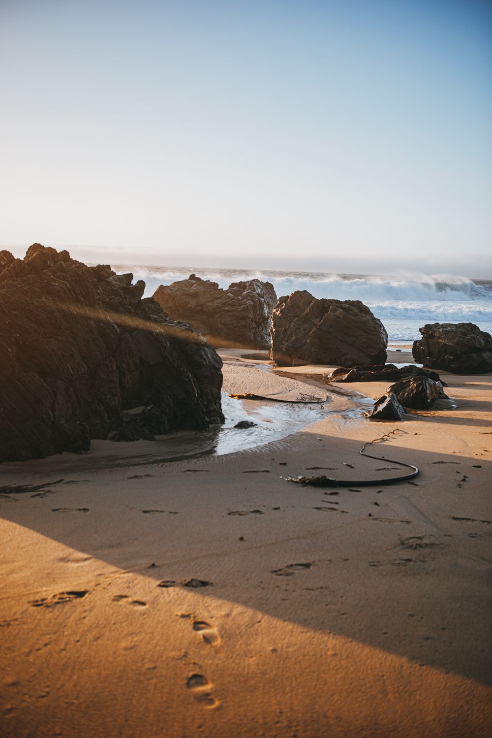 Rocky coastal beach landscape with dark volcanic rocks and crashing waves against golden sand at sunset.