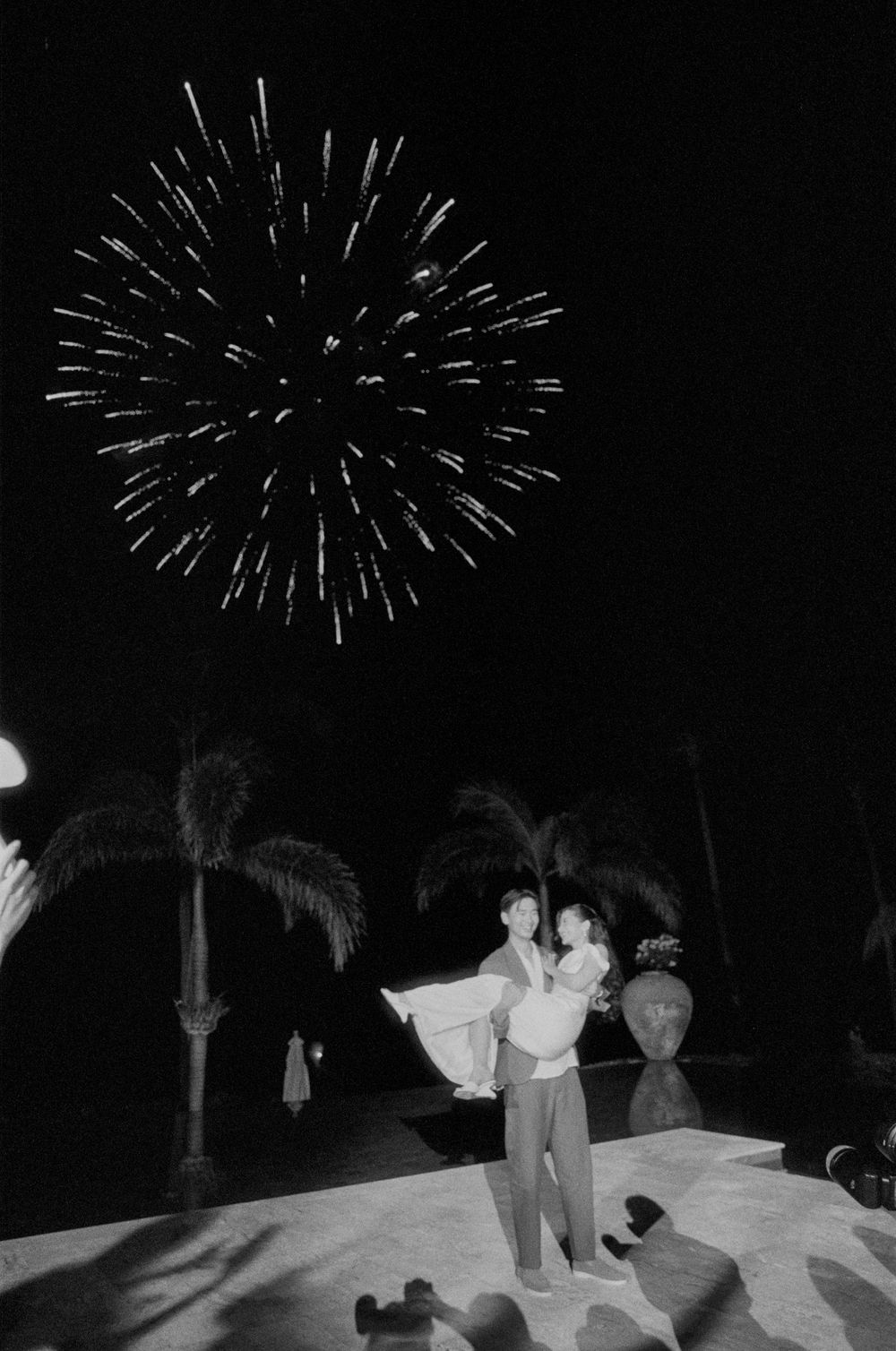 Fireworks light up the night sky over silhouettes of people and palm trees at an outdoor celebration.