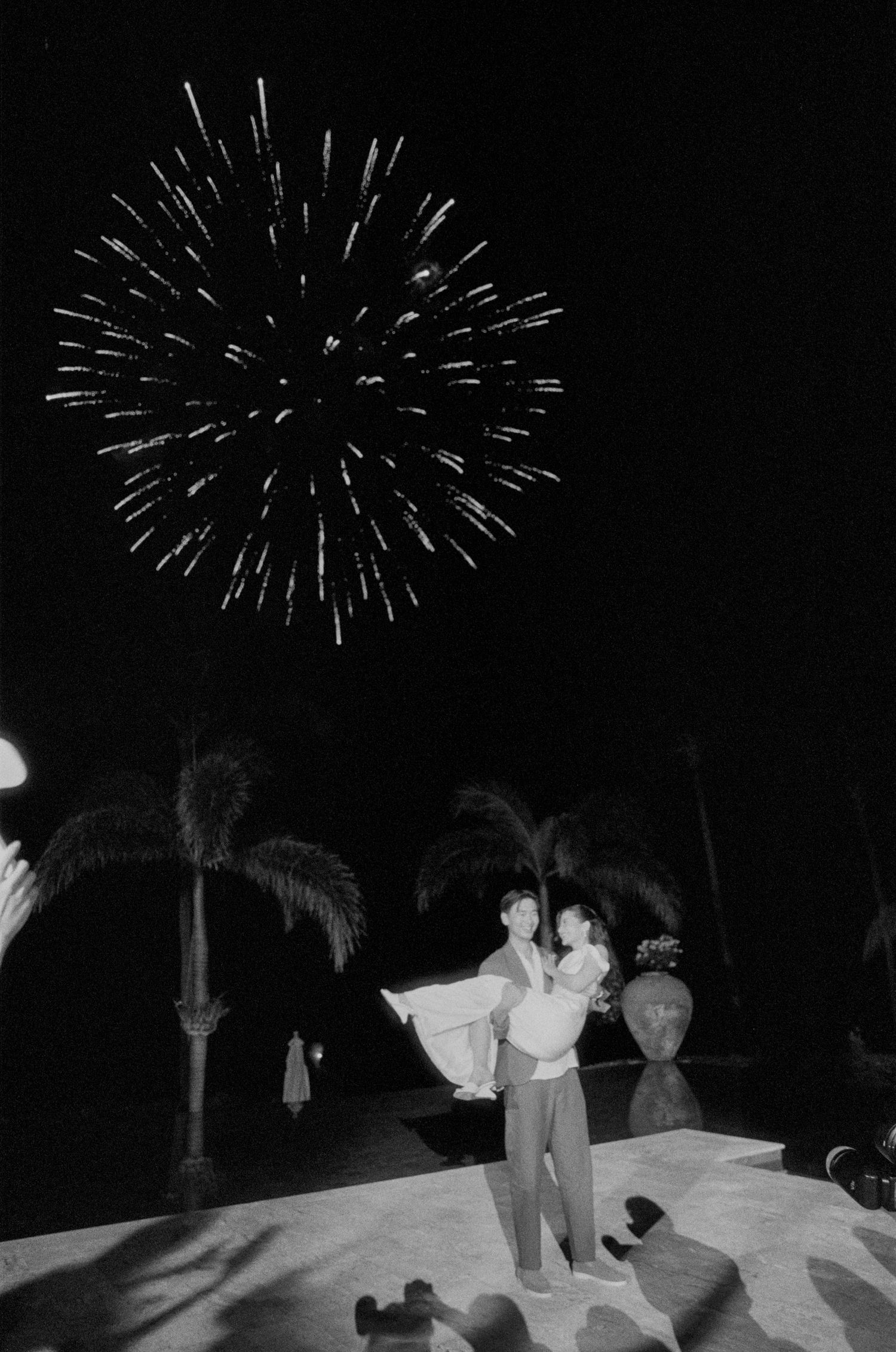Fireworks light up the night sky over silhouettes of people and palm trees at an outdoor celebration.